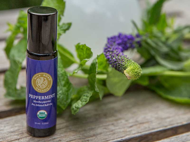 Silk Road organic peppermint essential oil roll-on bottle in front of a vase and flowers on a wooden tabletop