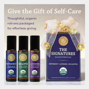 A gift set of three essential oil roll-on bottles with a matching branded box, shown on a light neutral background with promotional text. The set includes Peppermint, Eucalyptus, and Lavender bottles alongside a box labeled 'The Signatures' with a white ribbon and gold floral embellishments.