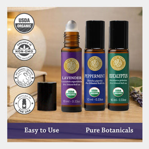 Three amber roll-on bottles with black caps are shown on a wooden surface with a blurred warm indoor background. The bottles have purple, blue, and green labels for Lavender, Peppermint, and Eucalyptus, with USDA Organic seal, non-gmo badge, cruelty-free, and bottled in the USA