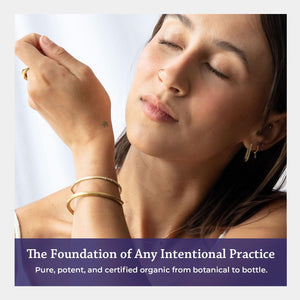 A woman with closed eyes and her hand near her face, wearing gold bracelets and earrings, with a purple text banner at the bottom reading 'The Foundation of Any Intentional Practice' and 'Pure, potent, and certified organic from botanical to bottle.'.