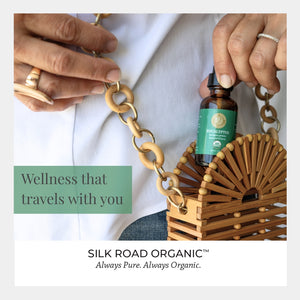 Silk Road Organic bottle held by a hand with a wooden bag and text 'Wellness that travels with you'.