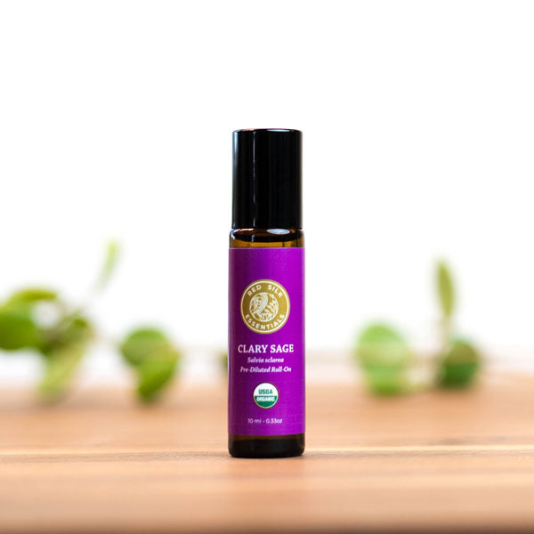 roll-on bottle of pre-diluted clary sage essential oil sits on a bright countertop with a. plant trailing in the background, bright magenta red silk essentials label