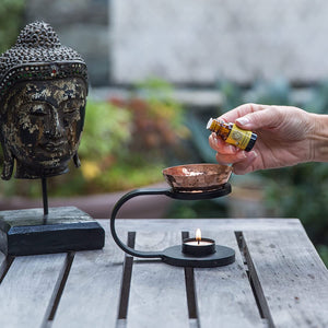 iron and copper essential oil burner sitting on a table outside, buddha head statue in bakground, person pouring in essential oils